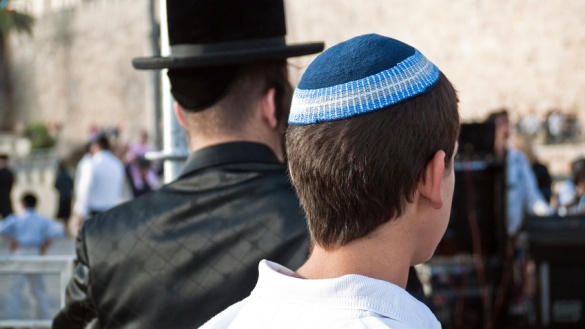 Mit dem Rücken zum Betrachter sieht man einen orthodoxen Juden in schwarzer Kleidung und mit einem schwarzem Hut. Daneben steht ein Jugendlicher mit einer blauen Kippa auf dem Kopf. Beide stehen vor der Klagemauer in Jerusalem.