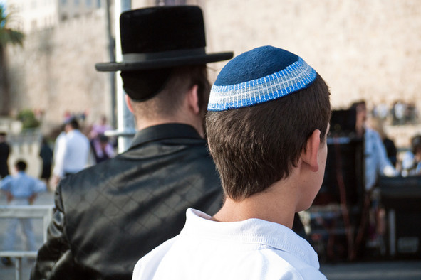 Mit dem Rücken zum Betrachter sieht man einen orthodoxen Juden in schwarzer Kleidung und mit einem schwarzem Hut. Daneben steht ein Jugendlicher mit einer blauen Kippa auf dem Kopf. Beide stehen vor der Klagemauer in Jerusalem.