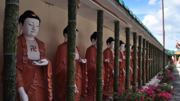 Blick auf eine Reihe Buddhastatuen in roten Gewändern hinter grünen schmalen Säulen.