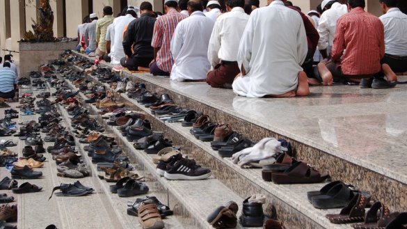 Auf einer Treppe vor einer Moschee stehen ganz viele Schuhe und man sieht betende Muslime.