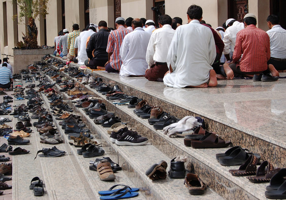 Auf einer Treppe vor einer Moschee stehen ganz viele Schuhe und man sieht betende Muslime.