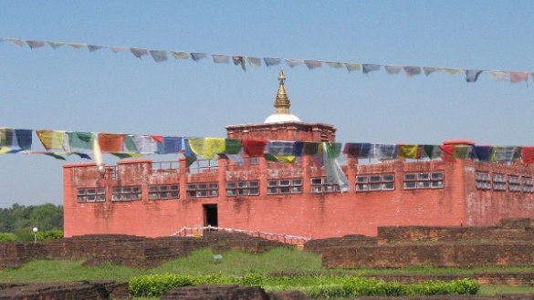 Ein roter, buddhistischer Tempel vor blauem Himmel mit aufgespannten Gebetsfahnen im Vordergrund. 