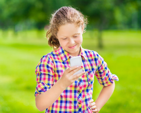 Ein Mädchen steht auf einer grüner Wiese mit einem Smartphone in der rechten Hand und tippt.
