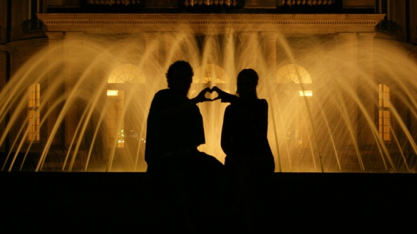 Silhouette eines Paares vor einem beleuchteten Brunnen bei Nacht, die mit den Händen ein Herz formen.