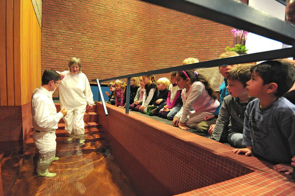 Eine erwachsene Frau in weißer Kleidung wird von einem Priester in weißer Kleidung im Wasserbecken der Freikirchlichen Gemeinde Hannover am Döhrener Turm zur Baptistin getauft. Kinder schauen zu.