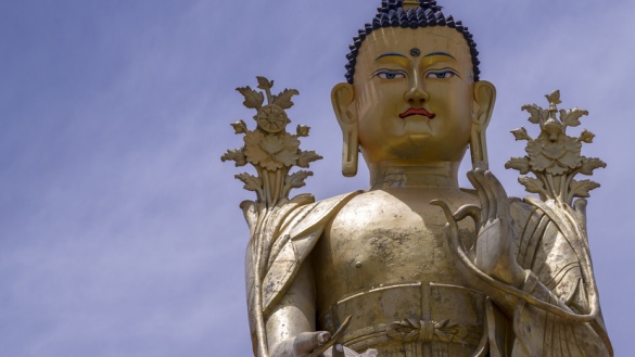 Goldene Buddha-Statue vor blauem Himmel dessen linke Hand mit Zeigefinger und Daumen das Rad der Lehre formt.
