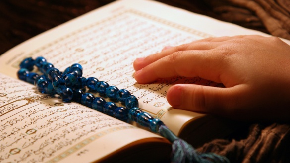 Eine blaue Gebetskette und eine Hand eines Kindes liegen auf einem aufgeschlagenen Koran.