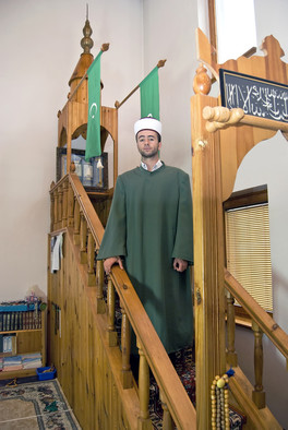 Ein Imam mit Kopfbedeckung und grüner Kleidung hält seine Ansprache auf der Treppe einer Gebetskanzel in einer Moschee.