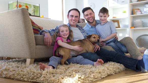 Ein homosexuelles männliches Paar sitzt mit Tochter, Sohn und Hund auf einem Teppich.
