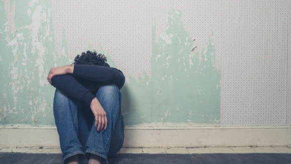 Trauriger junger Mann sitzt an einer Wand in einem leeren Raum und vergräbt den Kopf zwischen den Knien.