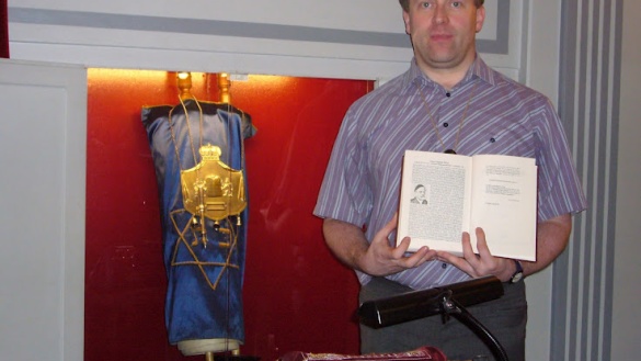 Eine Torarolle in einem Toraschrein, der mit rotem Samt ausgkleidet ist. Daneben steht ein Mann und hält ein aufgeschlagenes Buch vor sich.