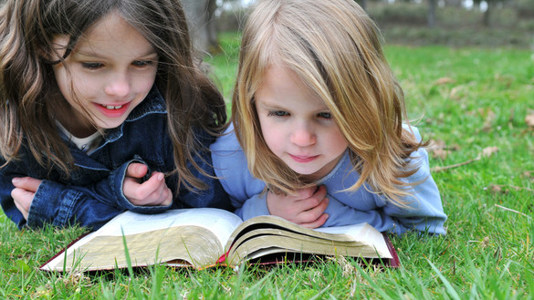 Zwei Mädchen liegen auf der grünen Wiese und lesen in einer aufgeschlagene Bibel