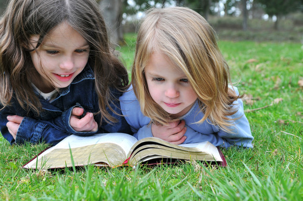 Zwei Mädchen liegen auf der grünen Wiese und lesen in einer aufgeschlagene Bibel