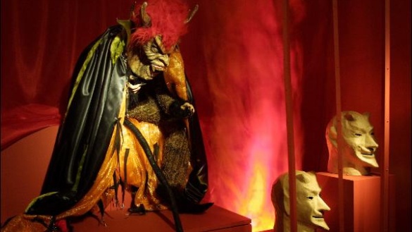 Eine Teufelsgestalt mit schwarzem Umhang, langem Schwanz, Hörnern und roten Haaren in einer Ausstellung.