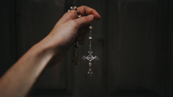 Hand hält Rosenkranz mit Kreuz vor einer dunklen Wand