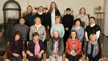 15 Frauen sitzen und stehen in drei Reihen hintereinander in einer Kirche und lächeln.