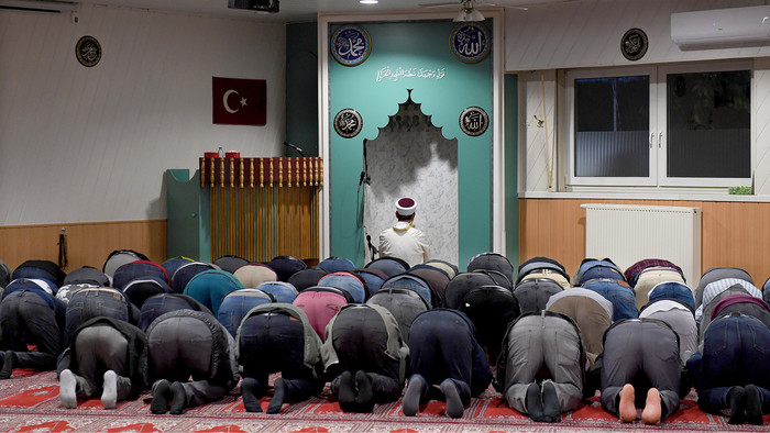 Mehrere Muslime neigen sich nach vorn zum Gebet, vor ihnen sitzt der Imam vor der Gebetsnische