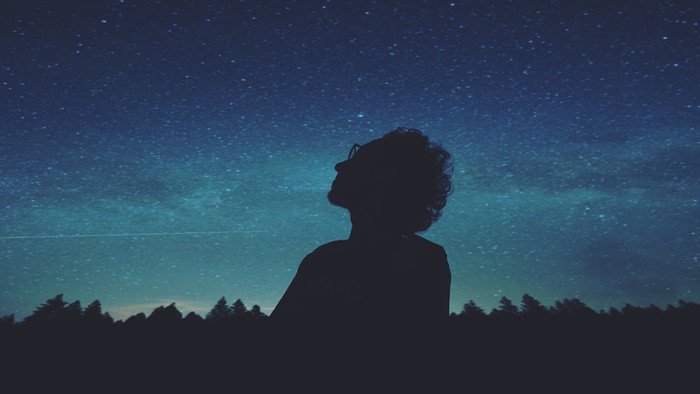Schwarze Silhouette eines Menschen mit Locken und Brille, der in einen Himmel in verschiedenen dunklen Blautönen, mit Sternen übersät, schaut.