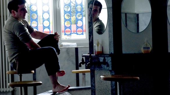 Ein Mann sitzt auf einem Hocker und wäscht seine Füße in einem Brunnen für rituelle Waschungen, rechts im Bild Spiegel über den Wasserhähnen und weitere Hocker